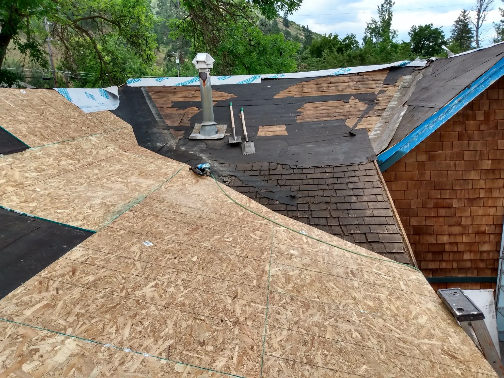 A house roof under renovation with sections of exposed plywood, partially removed shingles, and roofing tools; trees and a cloudy sky are visible in the background.