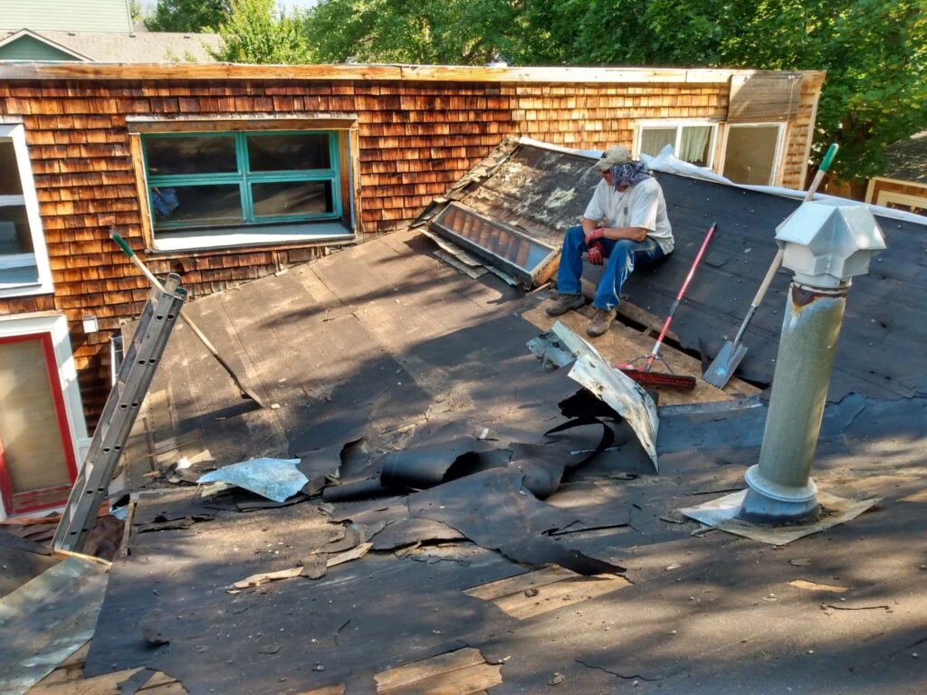 A person wearing a hat sits on a damaged shingle roof with torn and broken roofing materials, holding a tool. Ladders, a broom, and debris are visible around, with trees and a wooden-sided house in the background.