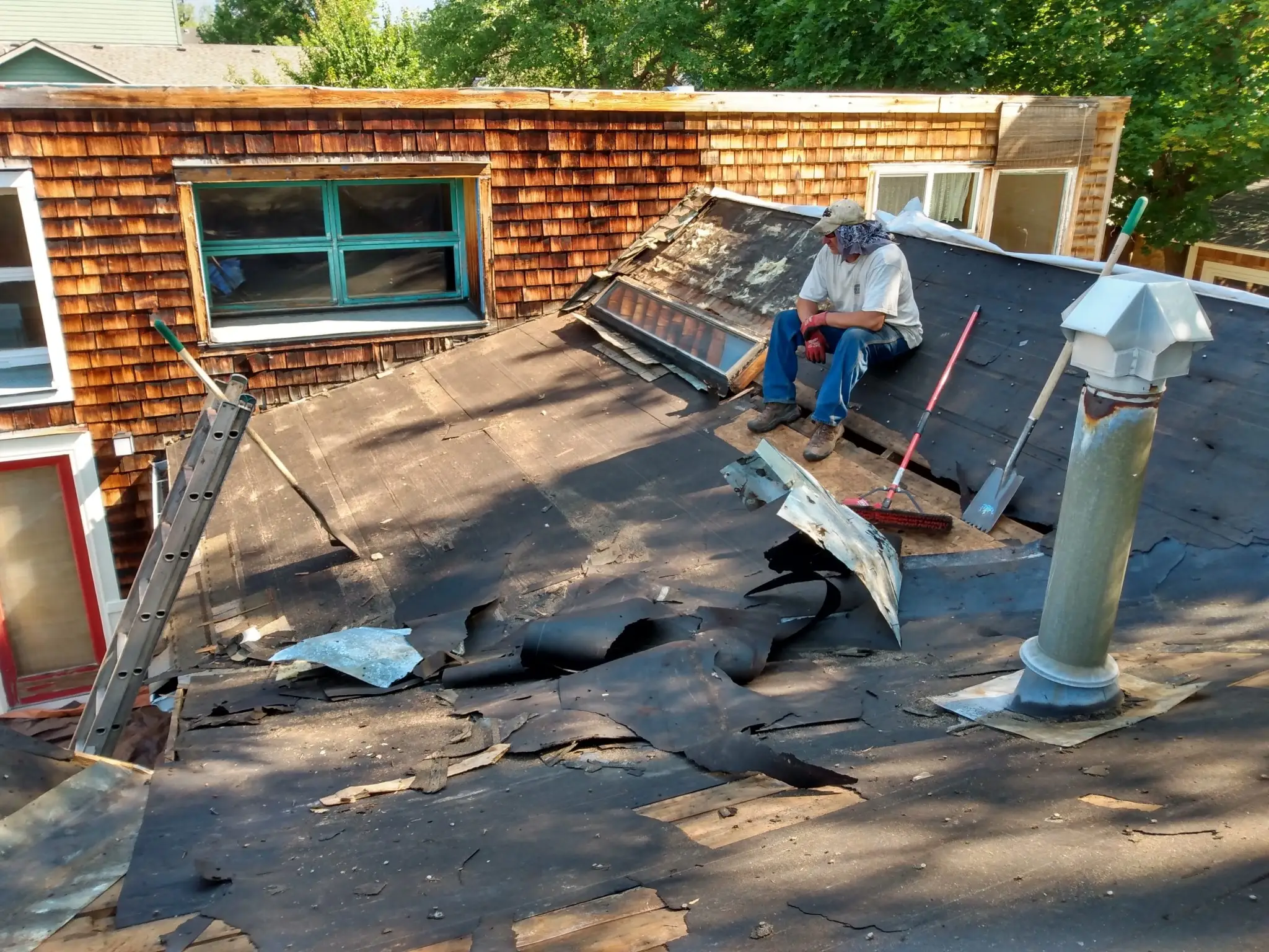 A person sits on a sloped roof removing old, damaged roofing material. Tools and debris are scattered around, with a ladder propped against the house. The house exterior is covered in wood shingles.