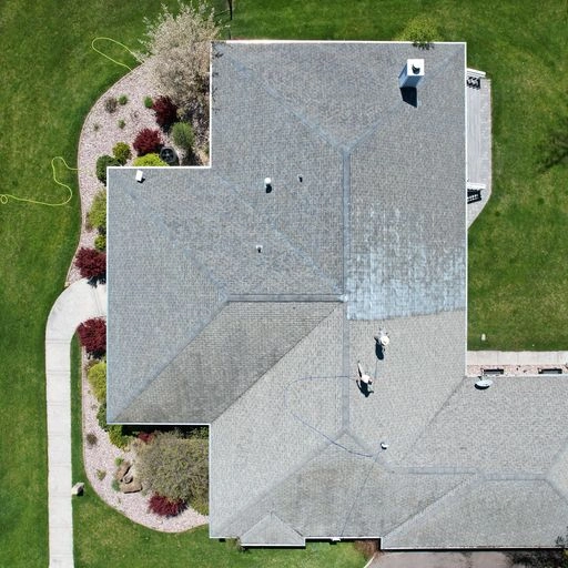Aerial view of a house with a gray roof, surrounded by green lawns, trees, landscaped garden beds, a sidewalk, and a yellow garden hose on the left side.