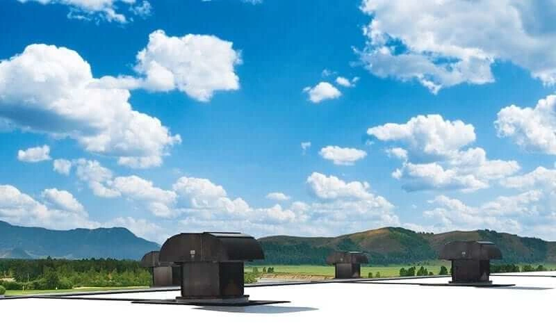 Rooftop with several black ventilation units under a bright blue sky filled with scattered clouds, with green hills and mountains in the background.