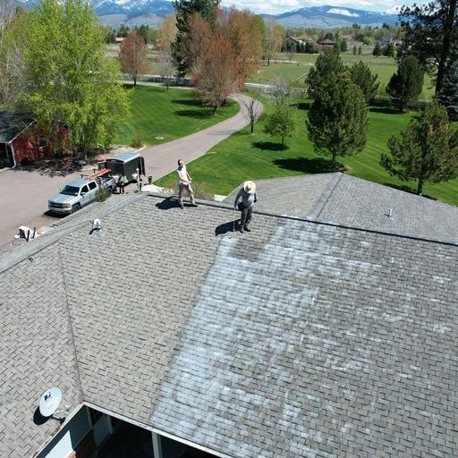Two workers stand on a shingled roof, cleaning or treating its surface. Vehicles and trees are visible below, and mountains can be seen in the distance under a partly cloudy sky.