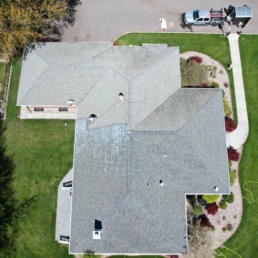 Aerial view of a house with a gray, multi-section shingle roof, surrounded by green lawn, pathways, and landscaped areas. A pickup truck and trailer are parked near the street at the top of the image.