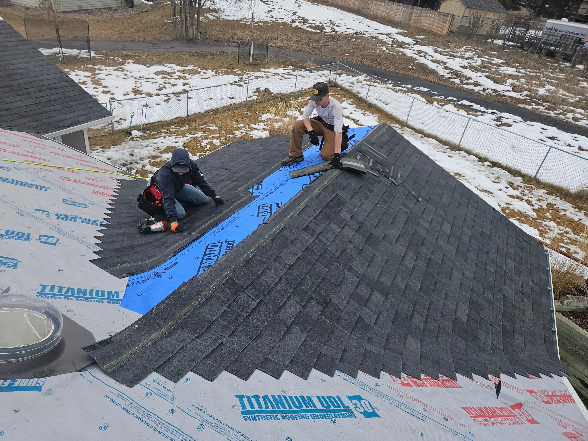 Two people are installing shingles on a house roof. One person kneels on the shingles while the other uses tools on roofing underlayment. Snow covers parts of the ground in the background.