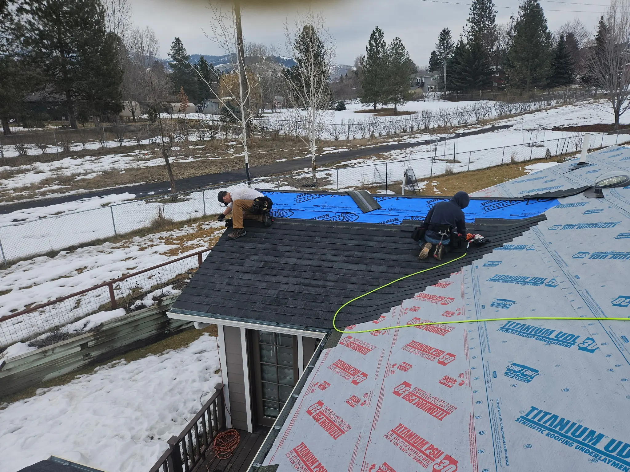 Two workers are installing shingles and underlayment on a snow-lined roof. Some sections are covered in blue and red synthetic underlayment, with trees and a partially snow-covered yard visible in the background.