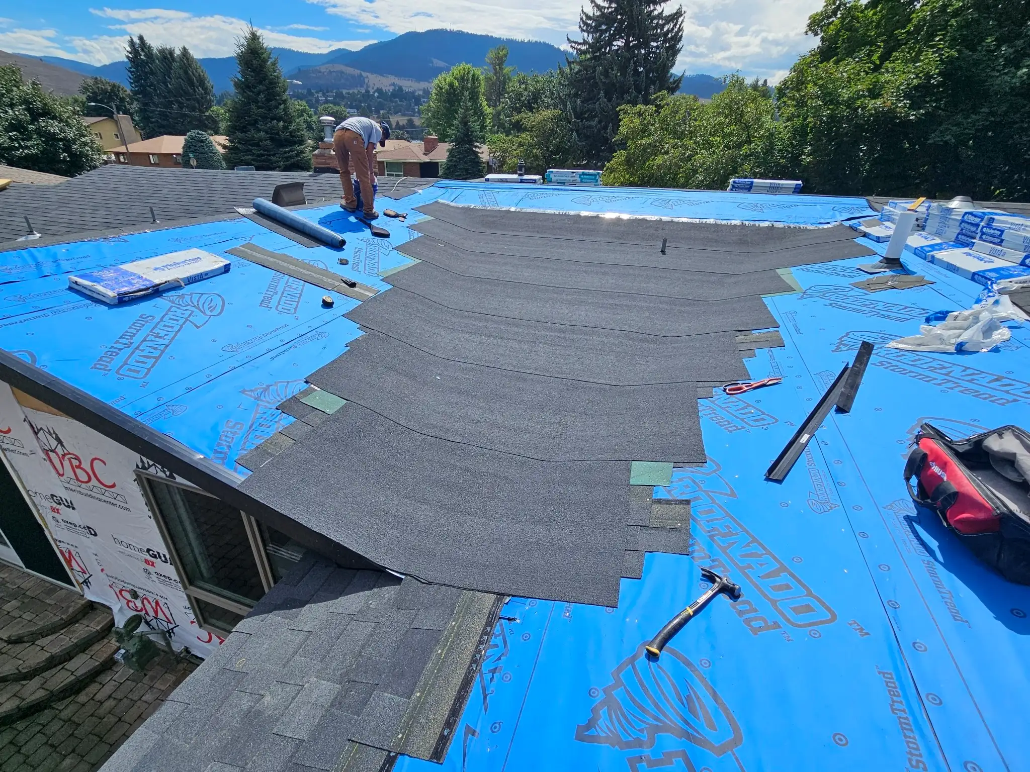 Two roofers work on a house covered with blue roofing underlayment, partially installing black shingles. Trees, houses, and mountains are visible in the background under a partly cloudy sky.