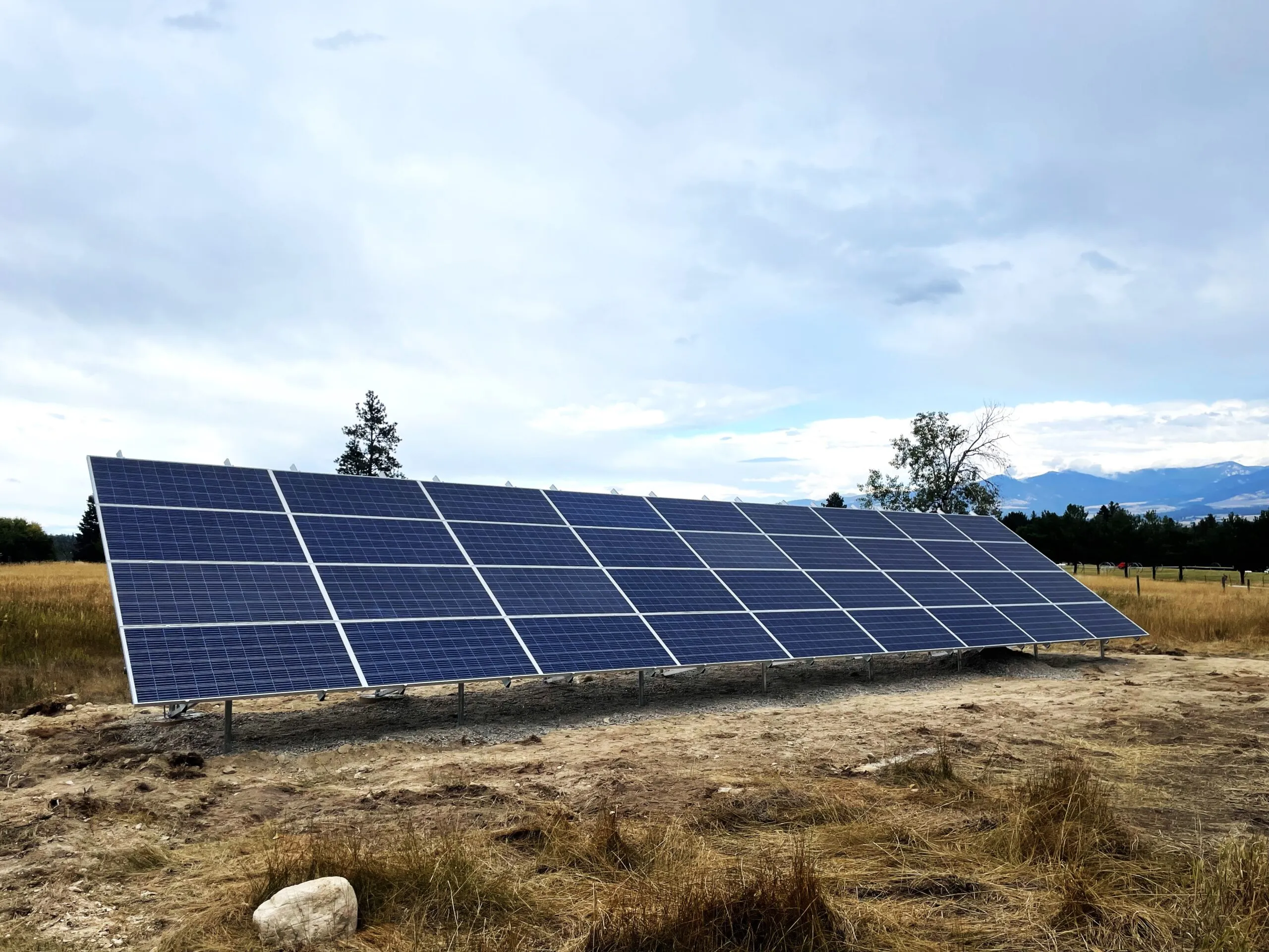 A row of solar panels stands on a dry, grassy field under a cloudy sky, with trees and distant mountains in the background.