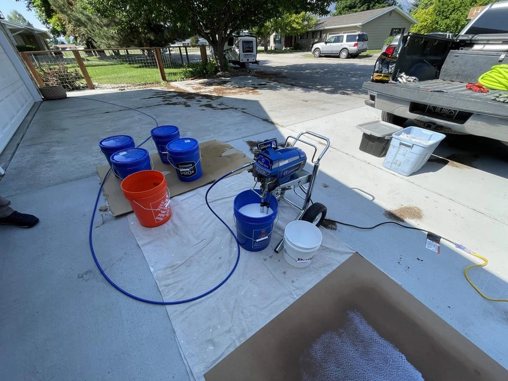 A driveway setup with several paint buckets, an airless paint sprayer on wheels, hoses, and drop cloths. A pickup truck is parked nearby, and trees and houses are visible in the background.