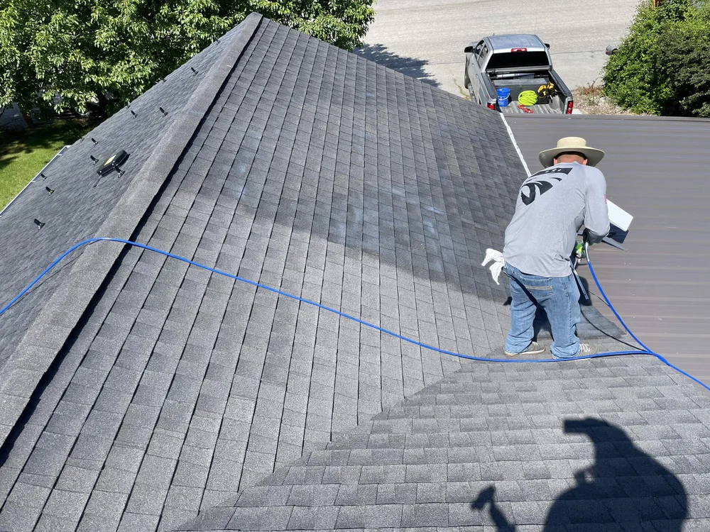 A person wearing a hat and long sleeves is cleaning a roof with a power washer, creating a visible contrast between the cleaned and uncleaned sections; a pickup truck is parked below.