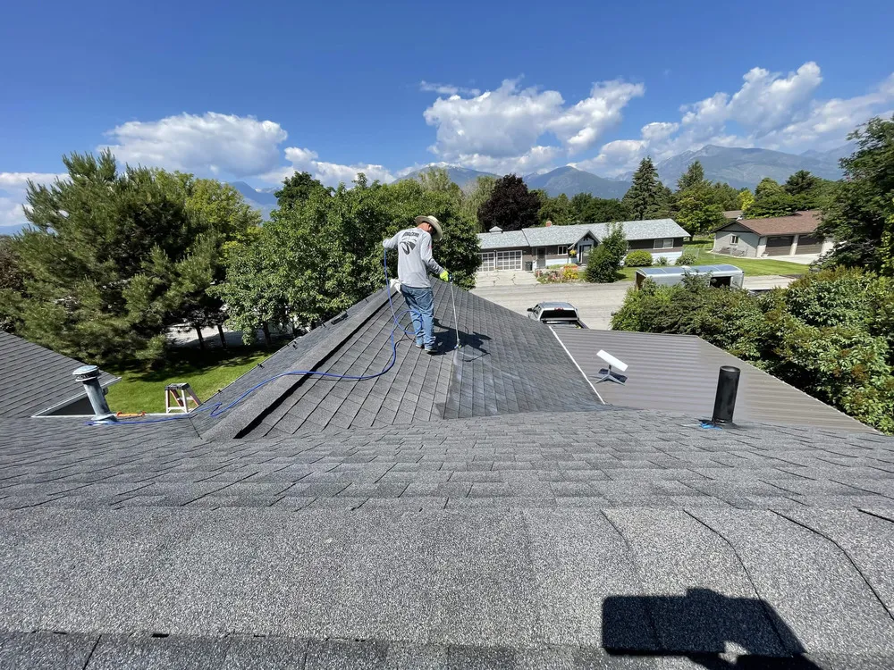 A person stands on the roof of a house, working with a paint roller and hose, applying a coating to the shingles on a sunny day with trees and mountains visible in the background.