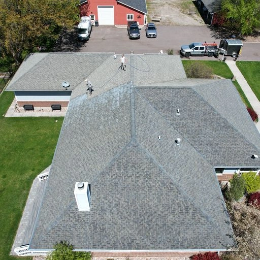 Aerial view of two people working on the roof of a large house with gray shingles, surrounded by green lawns, trees, parked vehicles, and red outbuildings in the background.