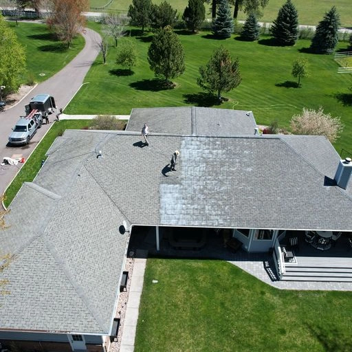 Aerial view of two workers cleaning a large gray roof on a suburban house, with cleaning equipment on the ground and green lawns and trees surrounding the property.