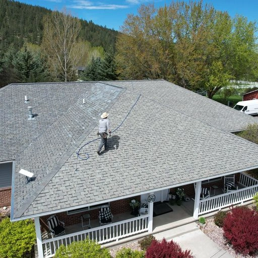 A person wearing safety gear is cleaning or spraying the sloped roof of a house with a hose. The house has a large front porch and is surrounded by trees and greenery under a blue sky.