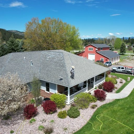 An aerial view of a house with a person working on the roof, surrounded by landscaped bushes and lawn, with a curved walkway, trees, and additional buildings and vehicles in the background on a sunny day.