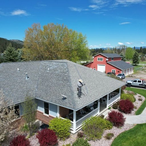 A person stands on the roof of a house using a hose or sprayer, surrounded by landscaped yard with shrubs, trees, and a curved stone path; a red barn and vehicles are visible in the background.