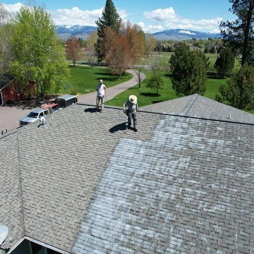 Two workers in hats clean or treat a house roof, with visible contrast between cleaned and uncleaned areas. Trees, vehicles, and mountains are visible in the background under a partly cloudy sky.