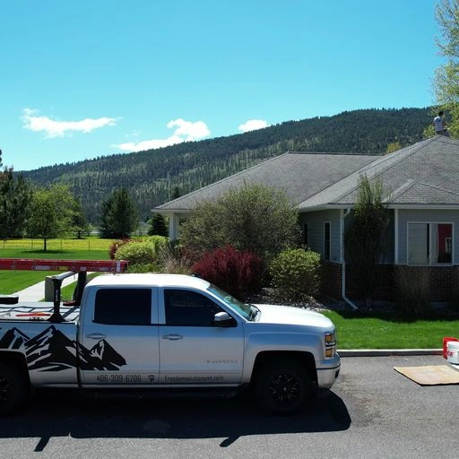A white pickup truck with a mountain graphic and business contact info is parked in front of a gray house with shrubs, trees, and a grassy yard, set against a backdrop of forested hills under a blue sky.