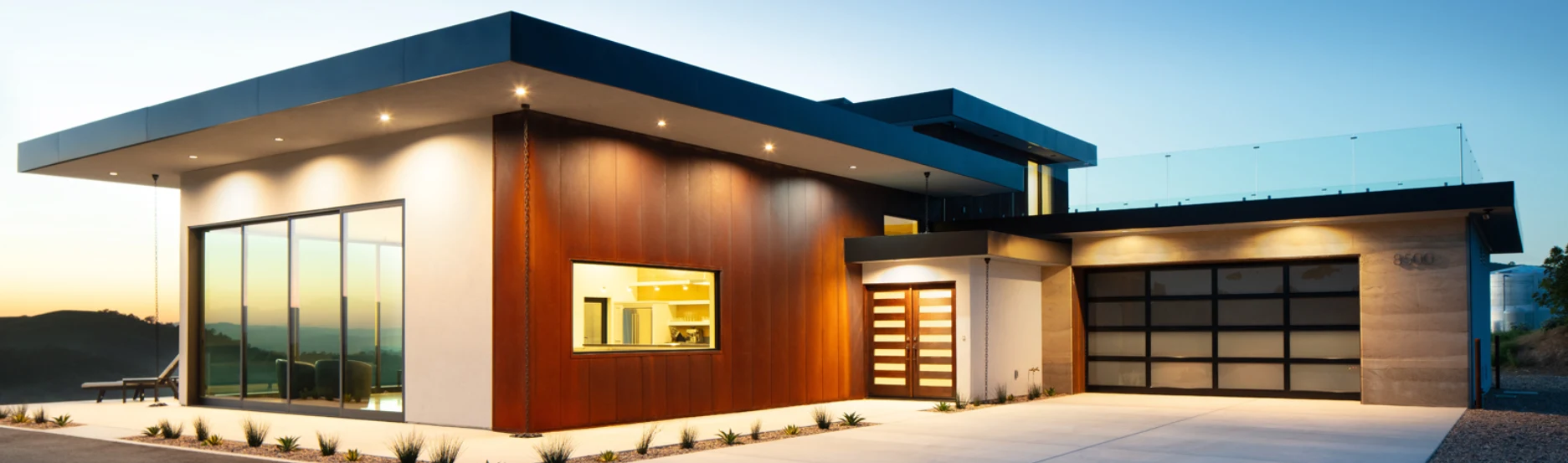 Modern single-story house at sunset with large windows, wood and concrete exterior, flat roof, illuminated entrance, and attached garage with a glass door. Minimalist landscaping lines the driveway.