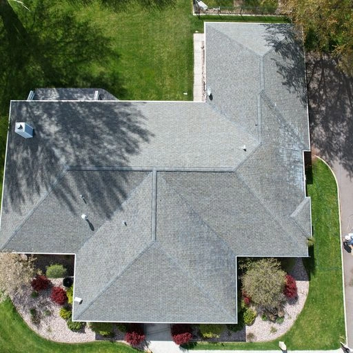 Aerial view of a house with a gray shingled roof, surrounded by green grass, trees casting shadows, a curved driveway, and landscaped garden beds.