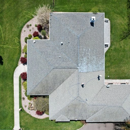 Aerial view of a house with a grey roof, surrounded by green lawn, curved sidewalks, and landscaped bushes and trees. The roof has various angles and vents visible.