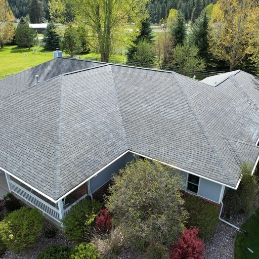 Aerial view of a house with a gray shingle roof, surrounded by trees, green lawn, and landscaped bushes in the yard.