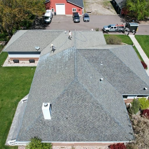 Aerial view of two workers inspecting or repairing the gray shingle roof of a large building surrounded by green grass, trees, and parked vehicles, with a red barn-like structure in the background.