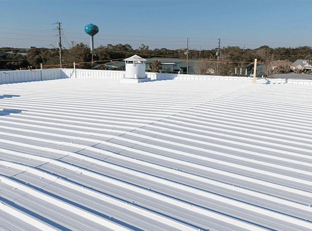 A white, reflective metal roof with raised seams is shown under clear blue sky, with trees, power lines, and a water tower visible in the background.