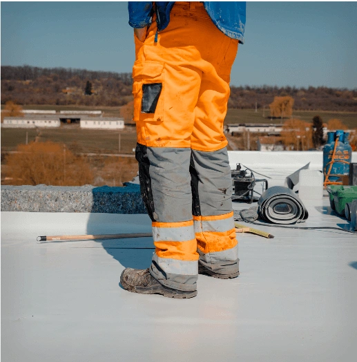 A person wearing bright orange work pants and boots stands on a rooftop, with roofing materials and tools nearby. The background shows a rural landscape with buildings, trees, and fields.