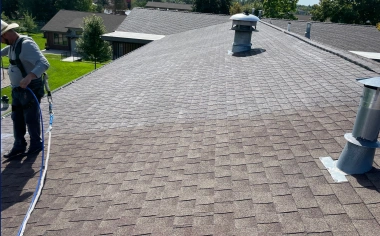 A person wearing a hat stands on a shingled roof, holding a hose or cable. There are two metal vents visible on the roof, with trees and buildings in the background under a sunny sky.