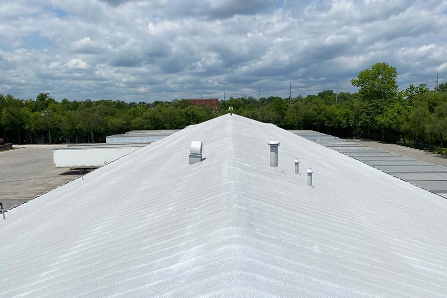View from the peak of a large, white metal roof with several vent pipes, overlooking a parking lot, trees, and cloudy sky in the background.