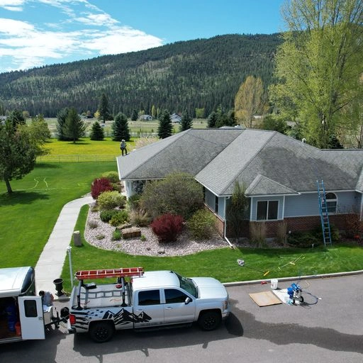 A white pickup truck with a ladder on top and a trailer attached is parked outside a suburban house with a gray roof. The house sits amid green lawns and trees, with forested hills in the background under a partly cloudy sky.