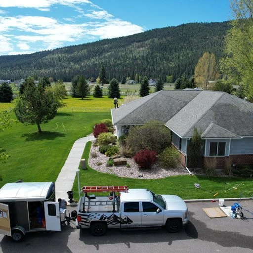 A white pickup truck with a trailer is parked on a driveway in front of a suburban house with a well-kept yard. Trees and mountains are visible in the background under a partly cloudy sky.