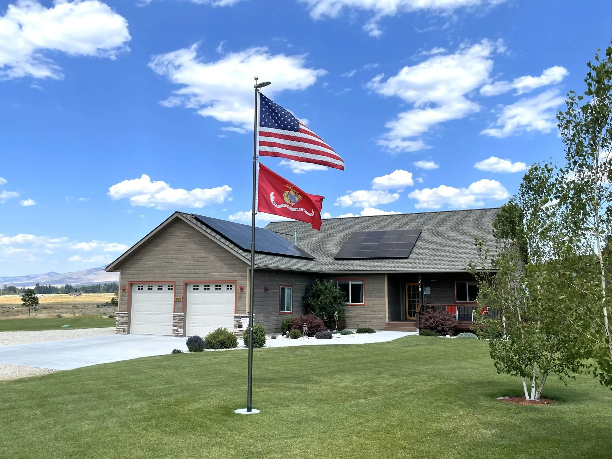 A house with solar panels on the roof sits behind a green lawn. Two flags, the American flag and a U.S. Marine Corps flag, are flying on a flagpole in front. It is a sunny day with a partly cloudy sky.