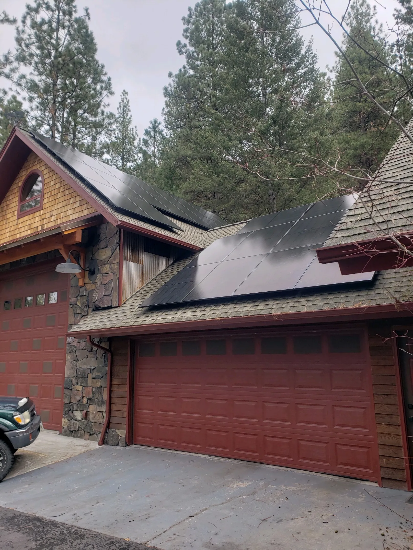 A house with two attached garages features rooftop solar panels. The exterior has stone and wood siding, surrounded by tall pine trees under a cloudy sky. A black vehicle is parked in front of the garages.