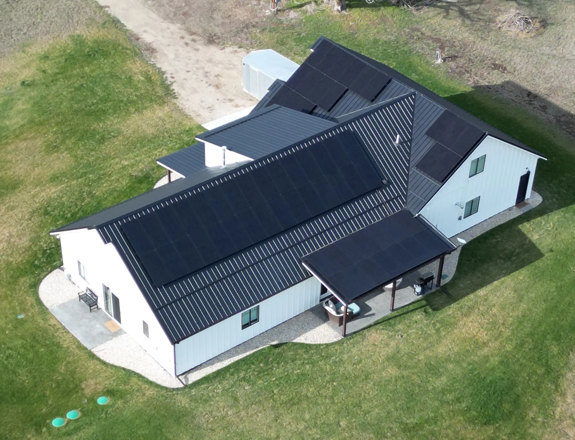 Aerial view of a white house with a black metal roof covered with solar panels. The home sits on a grassy area with a gravel pathway and a covered porch on one side.