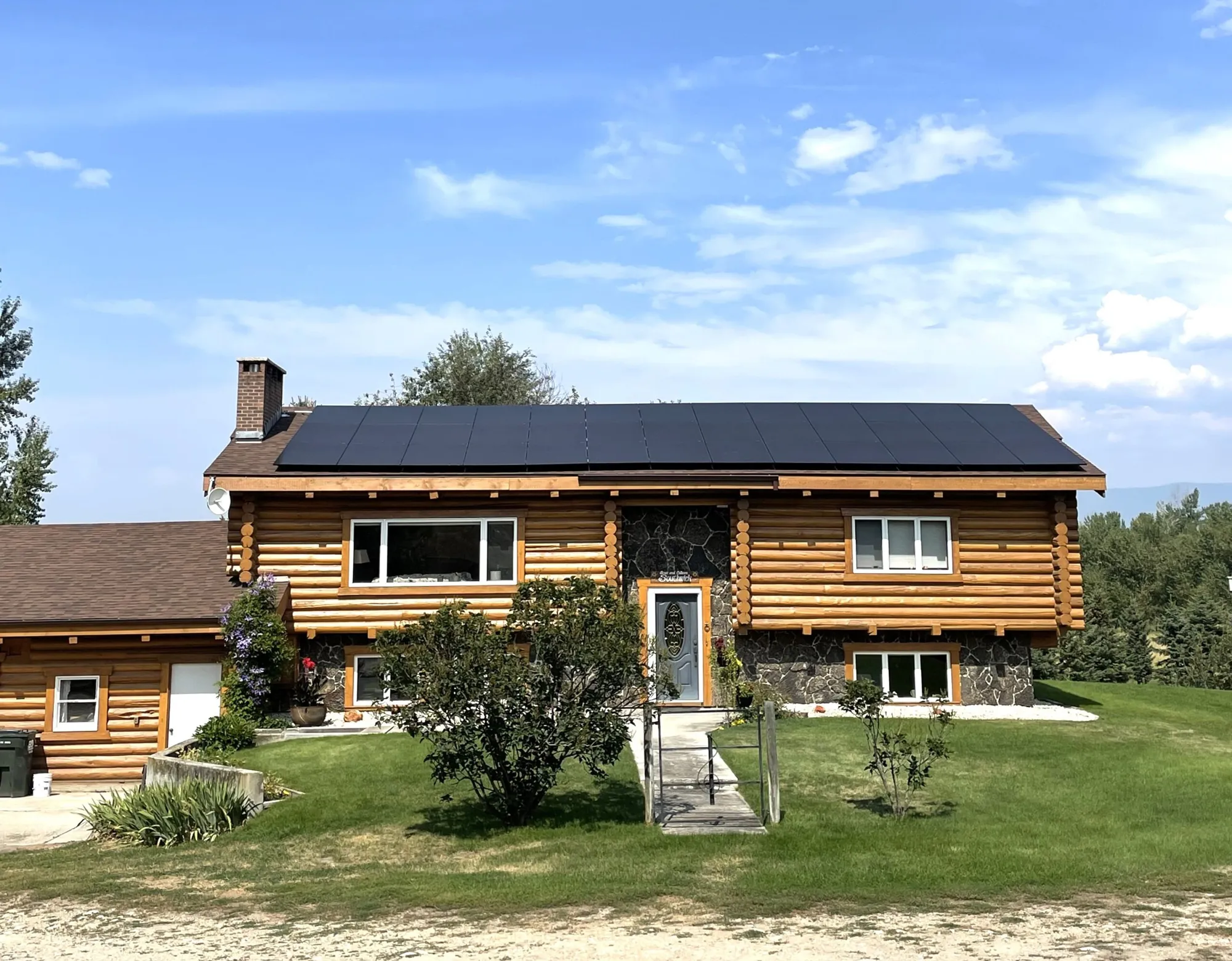 A two-story log cabin house with black solar panels on the roof, surrounded by green grass, bushes, and trees under a blue sky with scattered clouds.