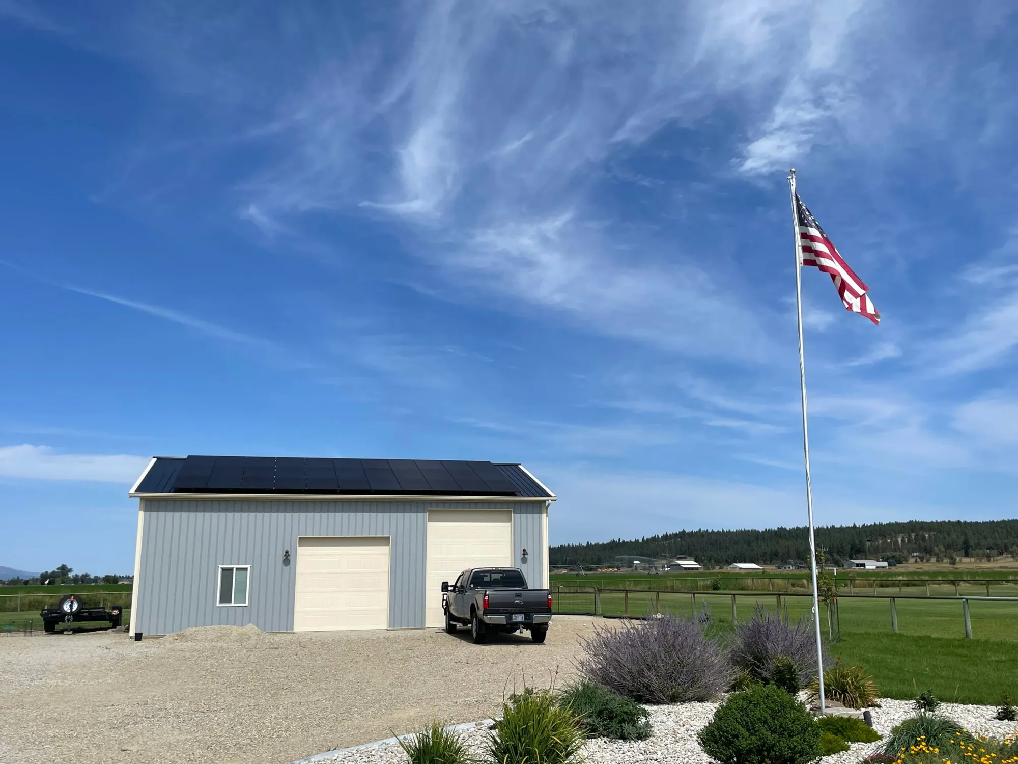 A gray metal garage with solar panels on its roof sits beside a parked black truck. An American flag on a tall pole waves in the foreground, with clear blue sky and scattered clouds above rural farmland.