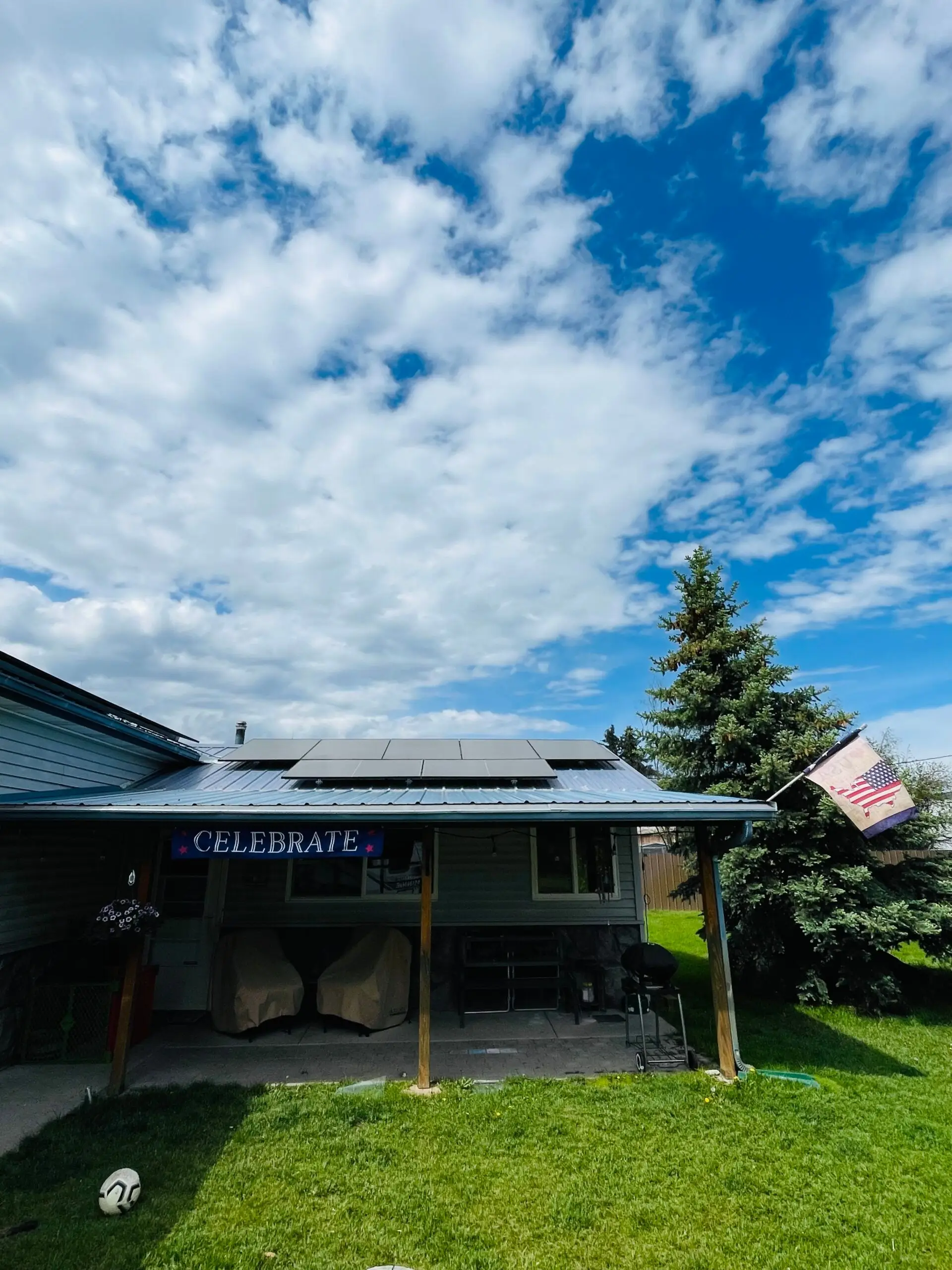 A house with solar panels on the roof, a Celebrate banner hanging on the porch, an American flag on a pole, green grass, and a partly cloudy blue sky overhead.