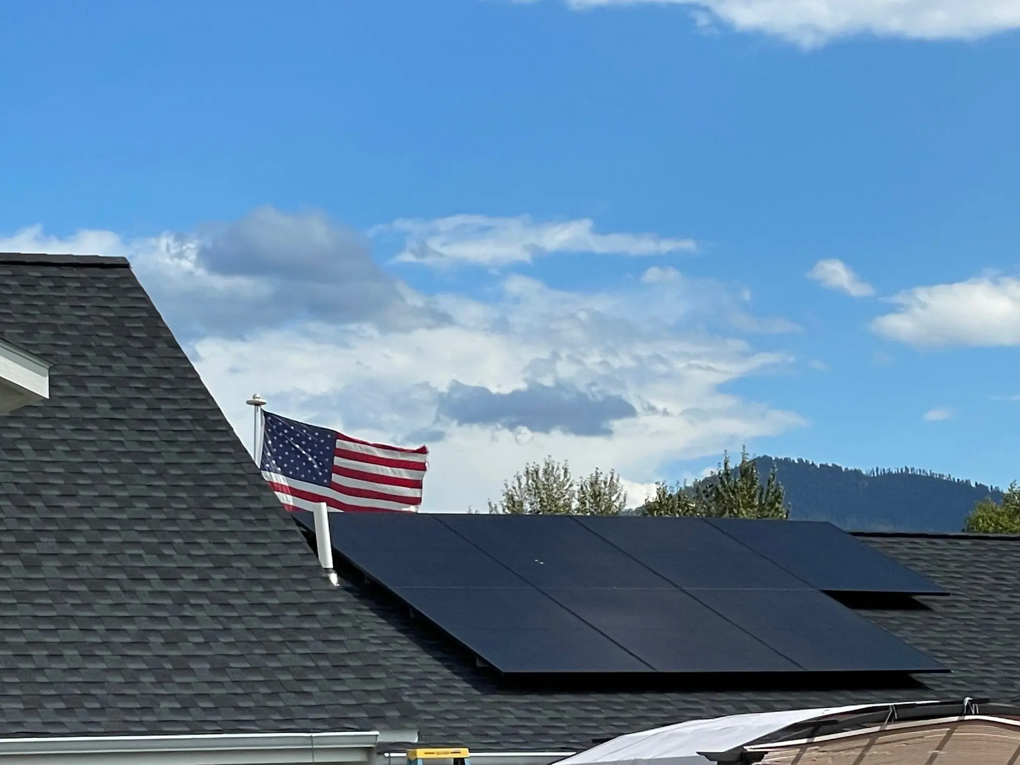 A row of black solar panels is mounted on a dark shingled roof. An American flag is displayed behind the house, with trees and a mountain visible under a blue sky with clouds in the background.