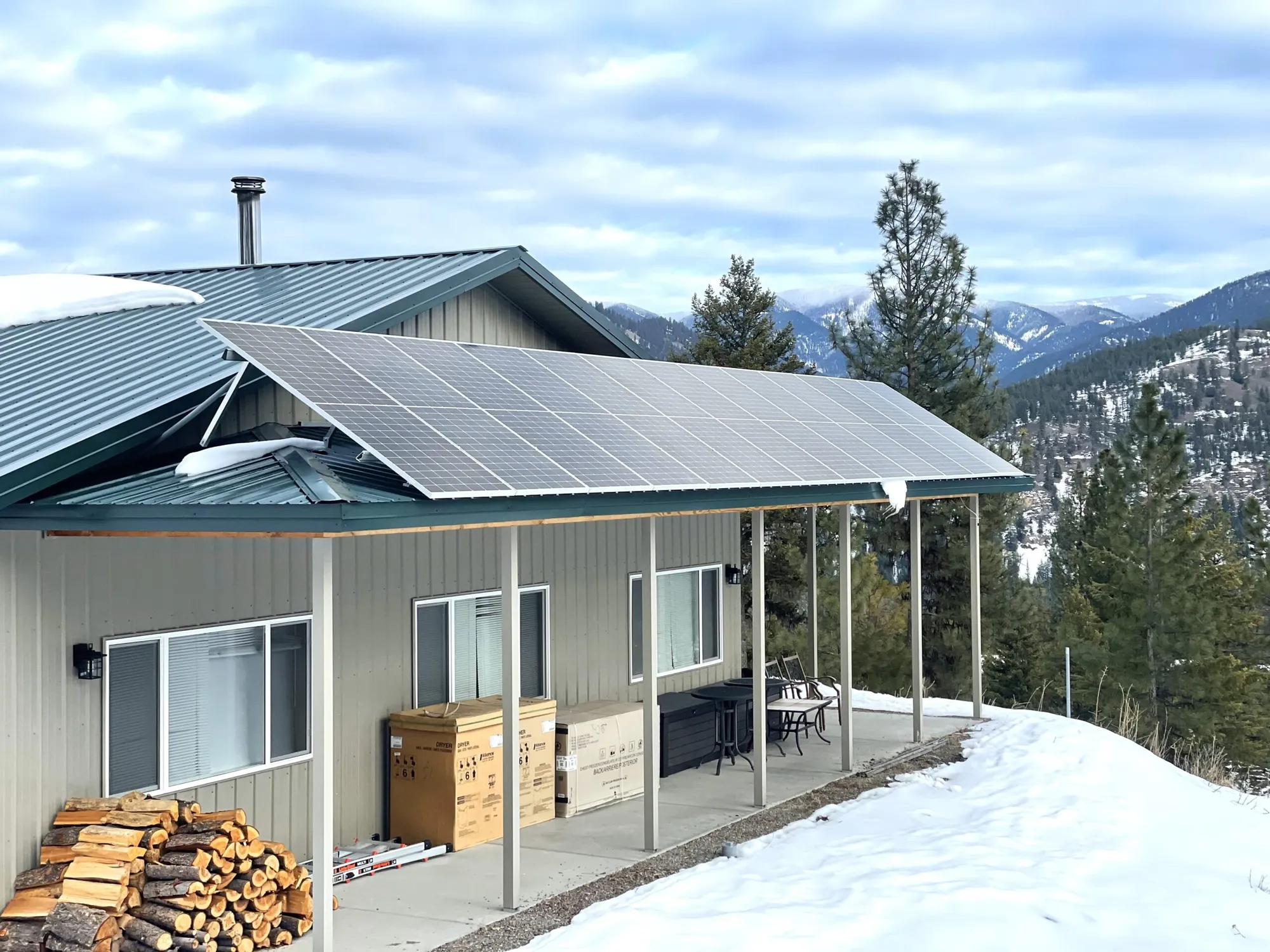 A metal-roofed house with solar panels installed on the roof, surrounded by snow, pine trees, and distant mountains under a cloudy sky. Firewood and cardboard boxes are stacked outside the house.