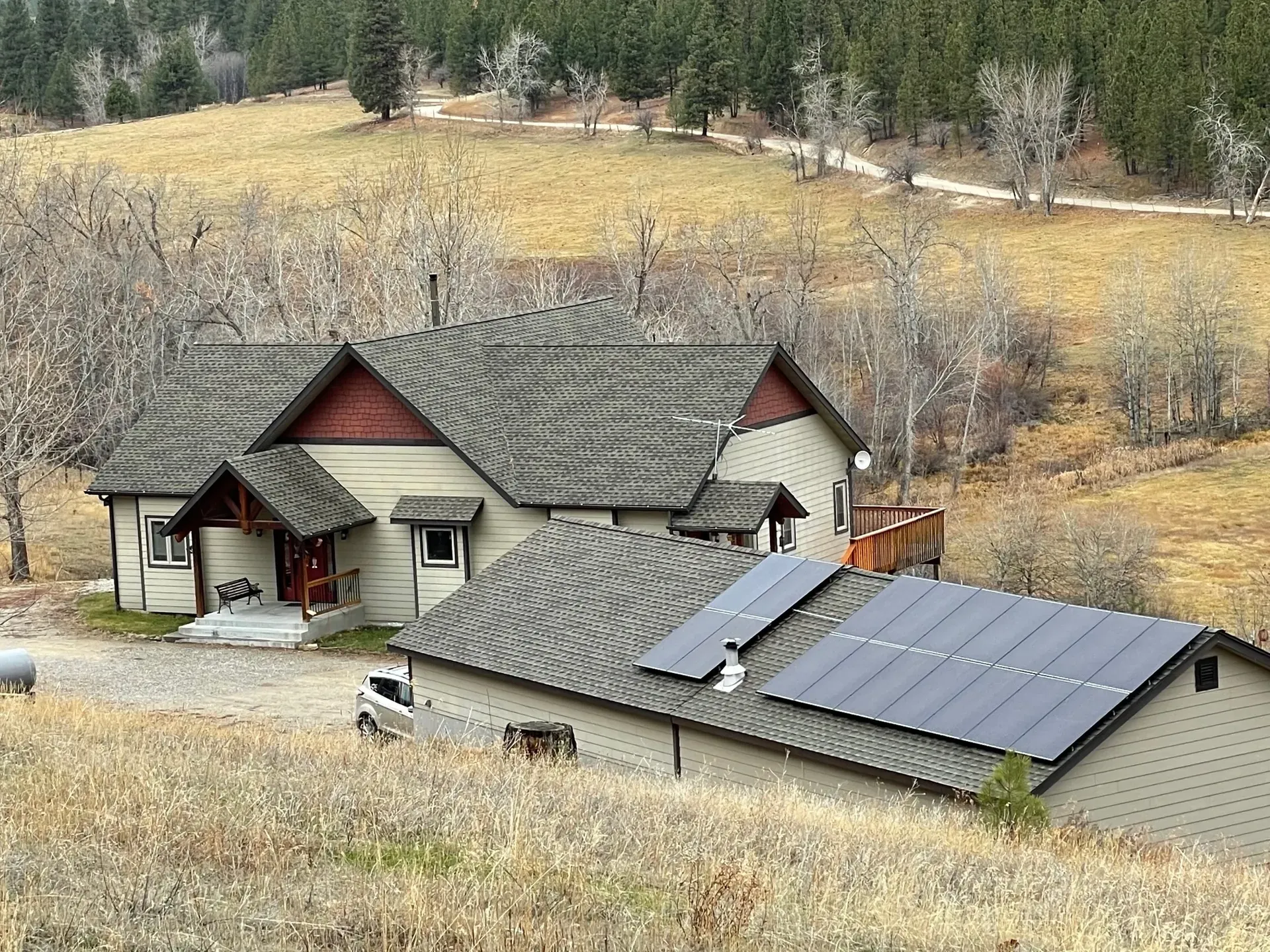 A house and detached garage with solar panels on the roof are set in a rural, hilly area with dry grass, leafless trees, and a forest in the background. A white vehicle is parked next to the garage.