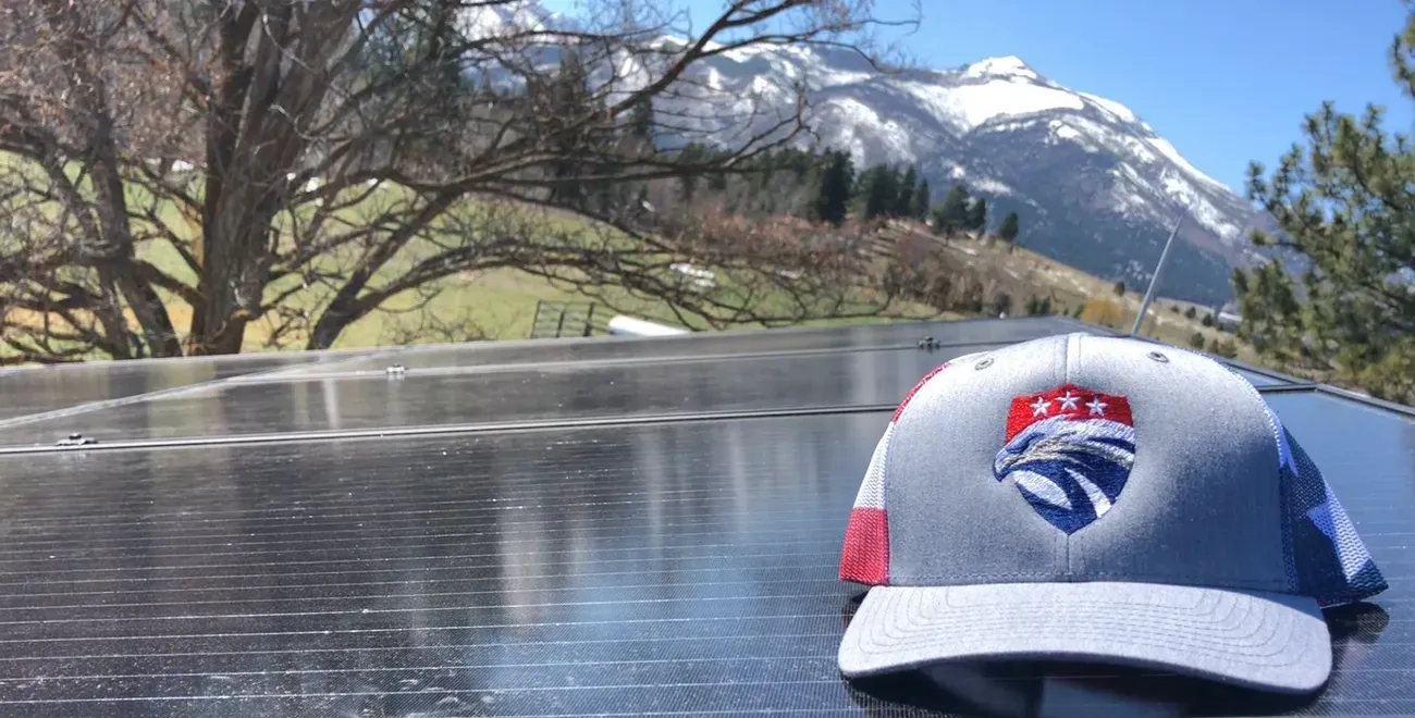 A gray and blue hat with a red, white, and blue emblem rests on a solar panel. In the background, there are green trees and fields, with snow-capped mountains under a clear blue sky.