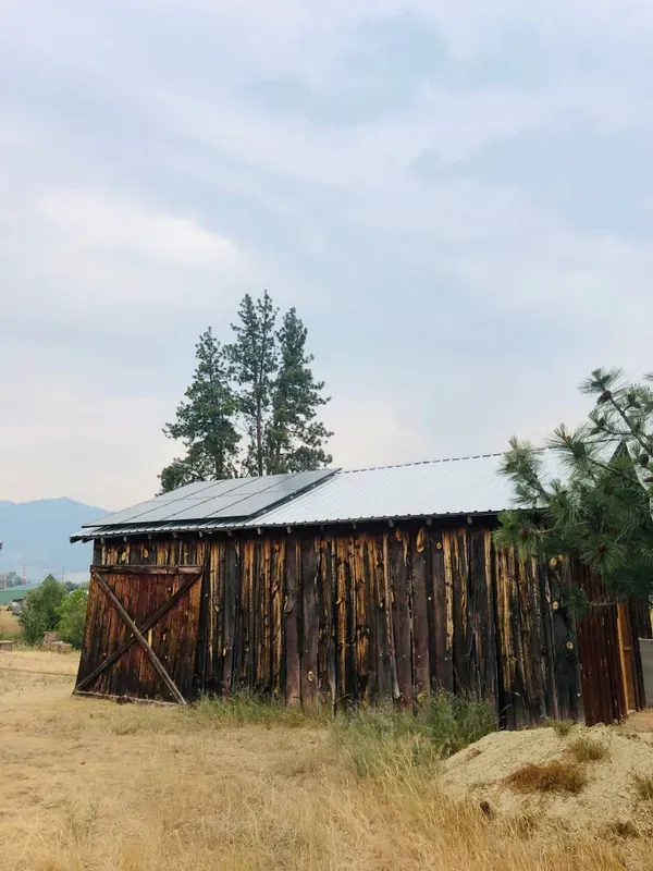 A rustic wooden barn with a tin roof stands on dry grass, surrounded by a few trees, under a cloudy sky with mountains visible in the background.