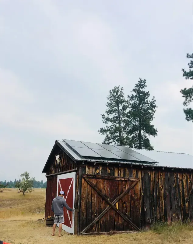 A person wearing a hat and shorts stands outside a weathered wooden shed with solar panels on the roof, opening a red and white door, surrounded by dry grass and tall trees under a cloudy sky.