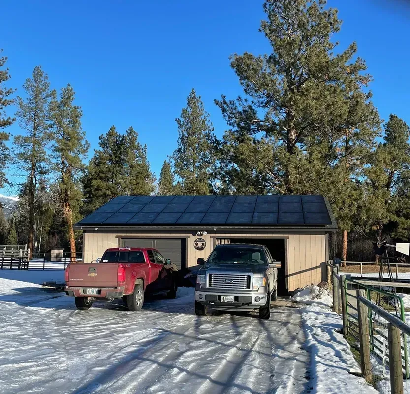 A snow-covered driveway leads to a tan garage with solar panels on the roof. A red pickup truck and a silver pickup truck are parked in front. Tall pine trees and a clear blue sky are in the background.