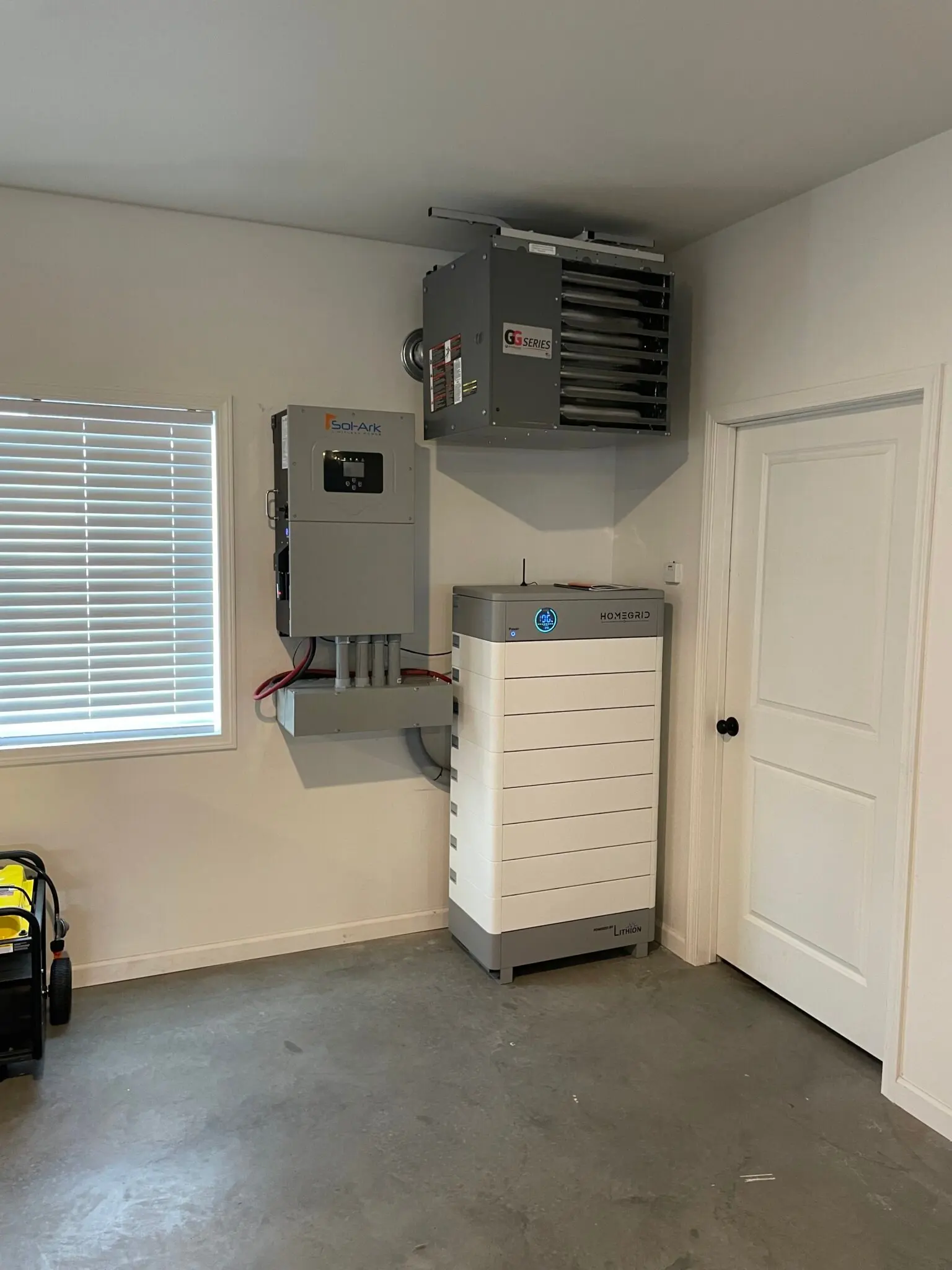 A home battery storage system is installed on the floor next to a wall-mounted electrical panel and a gray heater unit in a clean, organized garage with a closed white door and a small window with blinds.