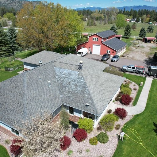 Aerial view of a house with a shingled roof, landscaped yard, and surrounding trees. A person stands on the roof. In the background are other buildings, vehicles, and distant mountains under a clear blue sky.