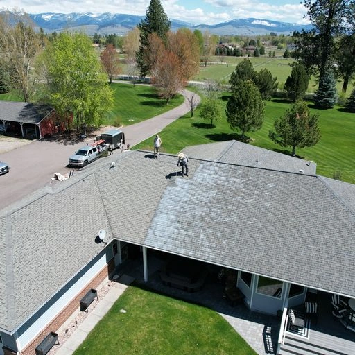 Aerial view of workers cleaning the large roof of a house surrounded by green lawns, trees, and mountains in the background under a partly cloudy sky.