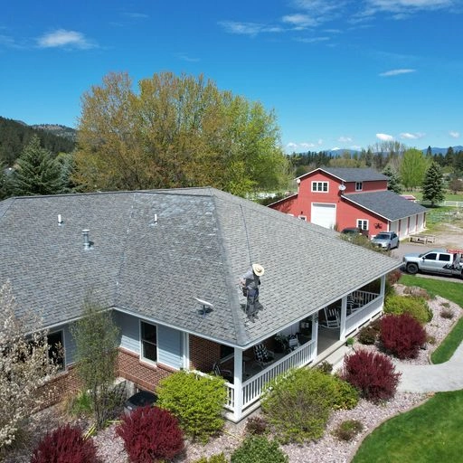 A worker in safety gear cleans or inspects the roof of a gray house with a wraparound porch, surrounded by landscaped bushes and trees. A red barn and white truck are visible in the background under a blue sky.