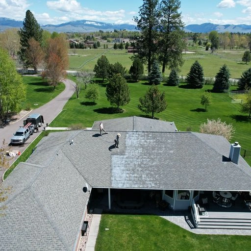Aerial view of two people cleaning or treating the roof of a suburban house with surrounding trees, grass, a driveway, a parked vehicle, and mountains visible in the distance.